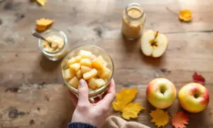 Main d'une personne tenant un bol de compote de pomme maison, entouré de pommes fraîches et de feuilles d'automne sur une table en bois.