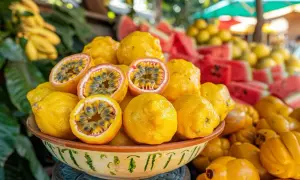 Un bol rempli de fruits jaunes, dont des fruits de la passion coupés révélant leur pulpe juteuse. Derrière, des pastèques et d'autres fruits en y.