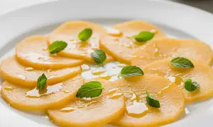 Carpaccio de melon frais, arrosé d'un filet de miel et garni de feuilles de menthe, sur une assiette blanche.