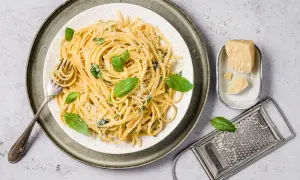 Assiette de pâtes au basilic frais et parmesan râpé, avec une fourchette prête à déguster. Un morceau de parmesan et une râpe complètent la scène.