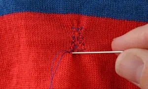 Main d'une personne utilisant une aiguille et du fil bleu pour comment recoudre un tissu déchiré sur un vêtement rayé rouge et bleu.