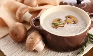 Un bol de soupe de champignons de Paris onctueuse, garnie de lamelles de champignons et de romarin. Des champignons frais entourent le plat.