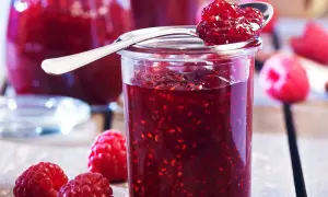 Un pot de confiture de framboise grand-mère, garni d'une cuillère pleine de cette délicieuse préparation. Des framboises fraîches entourent le pot.