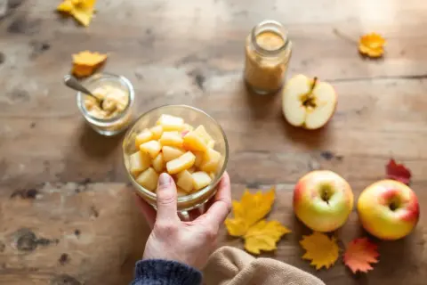 Main d'une personne tenant un bol de compote de pomme maison, entouré de pommes fraîches et de feuilles d'automne sur une table en bois.