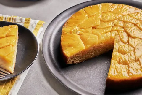 Un délicieux gâteau ananas renversé, avec une tranche servie sur une assiette noire. Les fruits caramélisés brillent sous la lumière.