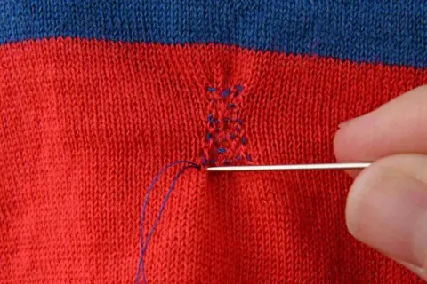Main d'une personne utilisant une aiguille et du fil bleu pour comment recoudre un tissu déchiré sur un vêtement rayé rouge et bleu.