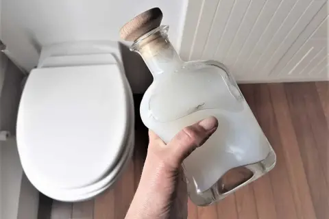 Main tenant une bouteille en verre remplie d'un produit wc maison, prêt à nettoyer la cuvette blanche.