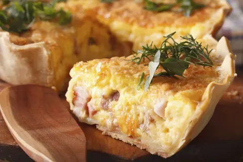 Une tranche de véritable quiche lorraine, garnie d'herbes fraîches, sur une planche en bois avec une cuillère.