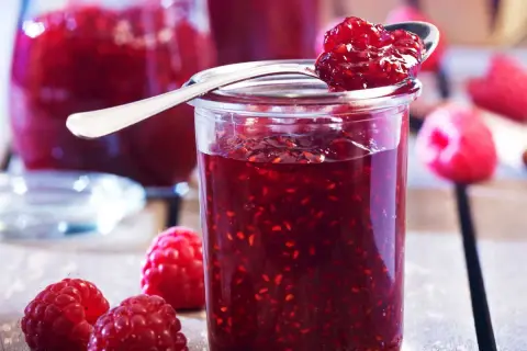 Un pot de confiture de framboise grand-mère, garni d'une cuillère pleine de cette délicieuse préparation. Des framboises fraîches entourent le pot.
