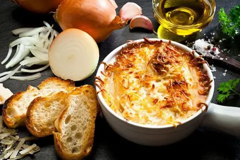 Une délicieuse soupe à l'oignon gratinée, servie avec des croûtons croustillants et du fromage fondu. Les ingrédients frais, comme les oignons et l'ail, sont disposés autour du plat.