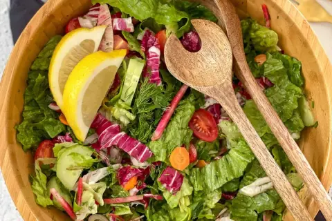Salade verte fraîche et colorée, parfaite pour une recette légère. Radis, concombre, tomates cerises et citron dans un saladier en bois avec des couverts.