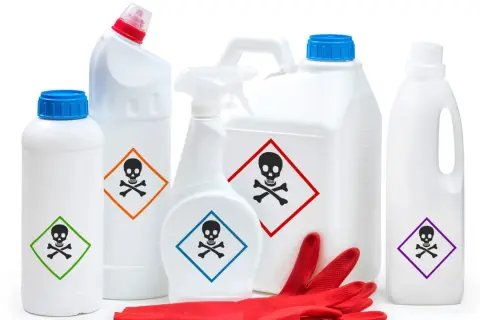 Plusieurs produits ménagers blancs, dont un pulvérisateur, portent un pictogramme toxique. Des gants rouges sont posés devant.