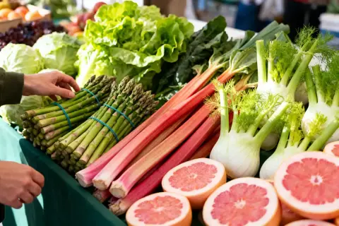 Un étal de marché débordant de fruits et légumes de saison : asperges, rhubarbe, fenouils, laitues et pamplemousses coupés.