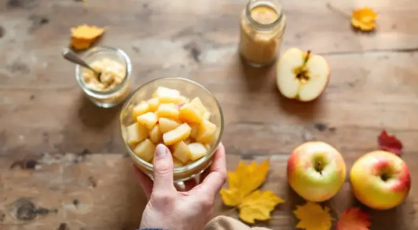 Main d'une personne tenant un bol de compote de pomme maison, entouré de pommes fraîches et de feuilles d'automne sur une table en bois.