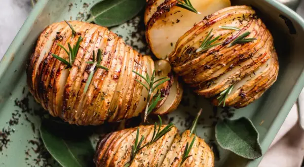 Pommes de terre Hasselback dorées et croustillantes, garnies de romarin frais et d'herbes, dans un plat vert.