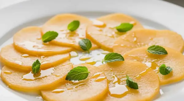 Carpaccio de melon frais, arrosé d'un filet de miel et garni de feuilles de menthe, sur une assiette blanche.