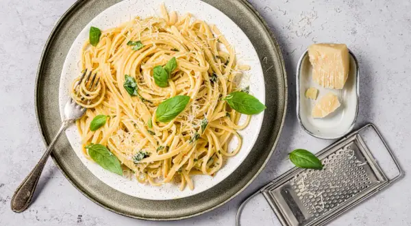 Assiette de pâtes au basilic frais et parmesan râpé, avec une fourchette prête à déguster. Un morceau de parmesan et une râpe complètent la scène.