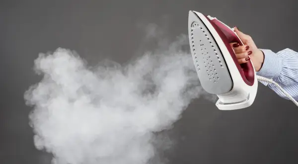 Une main tient un fer à repasser qui émet de la vapeur, suggérant l'action de détartrer le fer à repasser pour un entretien optimal.