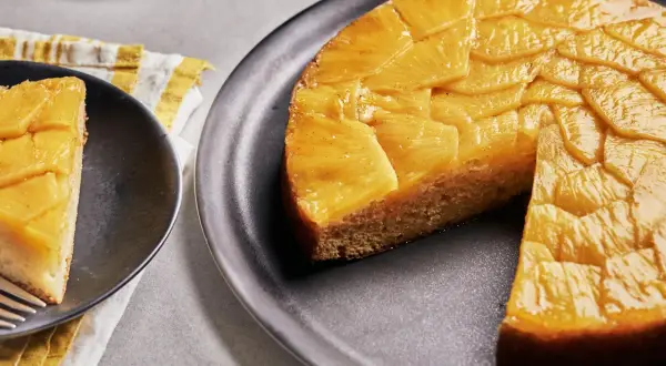 Un délicieux gâteau ananas renversé, avec une tranche servie sur une assiette noire. Les fruits caramélisés brillent sous la lumière.