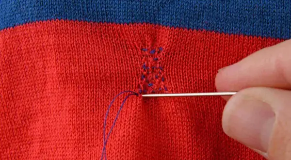 Main d'une personne utilisant une aiguille et du fil bleu pour comment recoudre un tissu déchiré sur un vêtement rayé rouge et bleu.