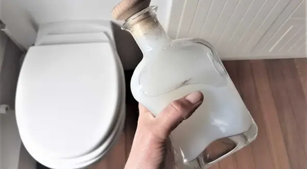 Main tenant une bouteille en verre remplie d'un produit wc maison, prêt à nettoyer la cuvette blanche.