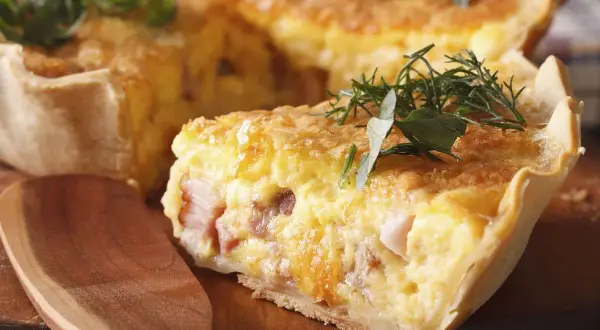 Une tranche de véritable quiche lorraine, garnie d'herbes fraîches, sur une planche en bois avec une cuillère.