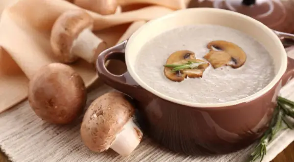 Un bol de soupe de champignons de Paris onctueuse, garnie de lamelles de champignons et de romarin. Des champignons frais entourent le plat.