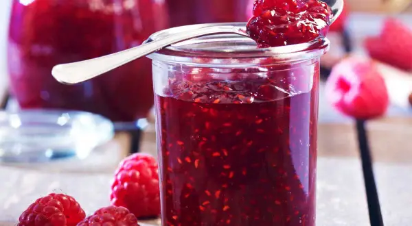 Un pot de confiture de framboise grand-mère, garni d'une cuillère pleine de cette délicieuse préparation. Des framboises fraîches entourent le pot.
