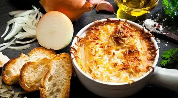Une délicieuse soupe à l'oignon gratinée, servie avec des croûtons croustillants et du fromage fondu. Les ingrédients frais, comme les oignons et l'ail, sont disposés autour du plat.