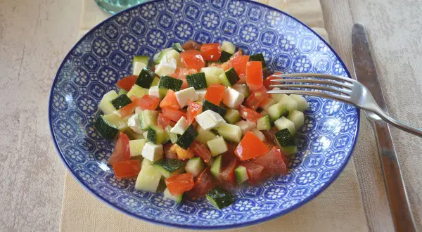 Salade de courgettes à la grecque, fraîche et colorée, avec des dés de tomates, courgettes et feta dans une assiette bleue.