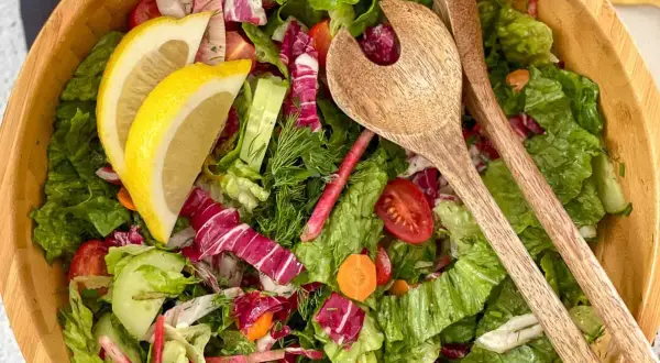 Salade verte fraîche et colorée, parfaite pour une recette légère. Radis, concombre, tomates cerises et citron dans un saladier en bois avec des couverts.