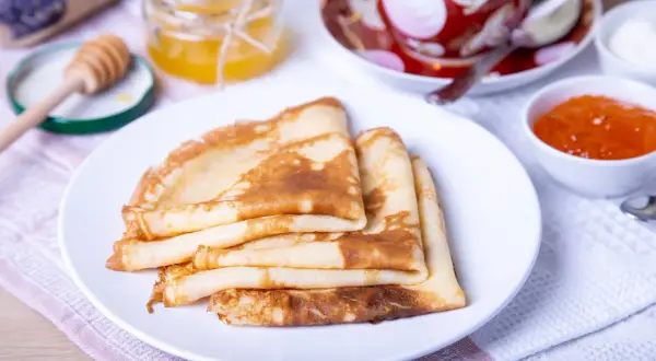 Pile de crêpes dorées, prêtes pour la préparation de la pâte à crêpes. Accompagnées de miel, confiture et d'une tasse de thé.