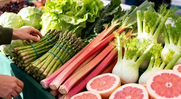 Un étal de marché débordant de fruits et légumes de saison : asperges, rhubarbe, fenouils, laitues et pamplemousses coupés.