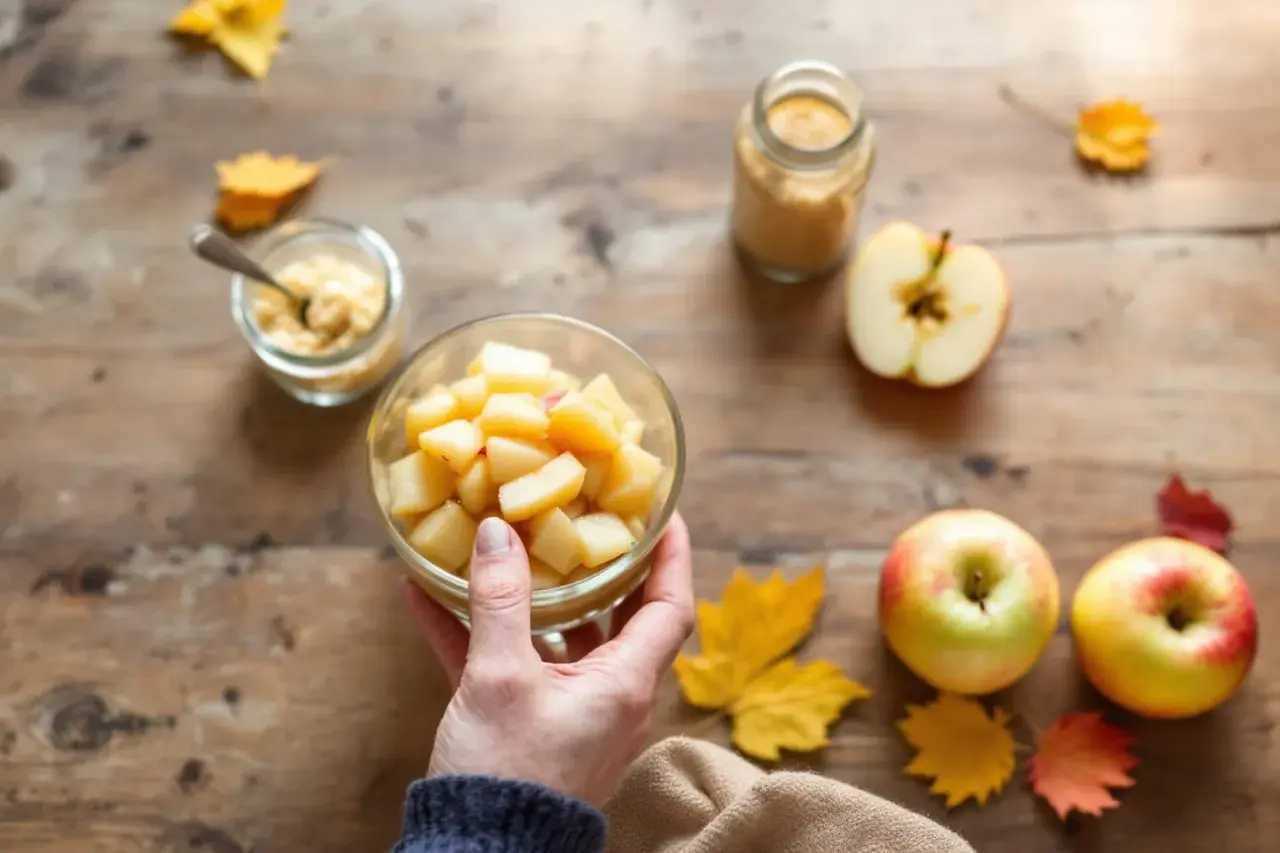 Main d'une personne tenant un bol de compote de pomme maison, entouré de pommes fraîches et de feuilles d'automne sur une table en bois.