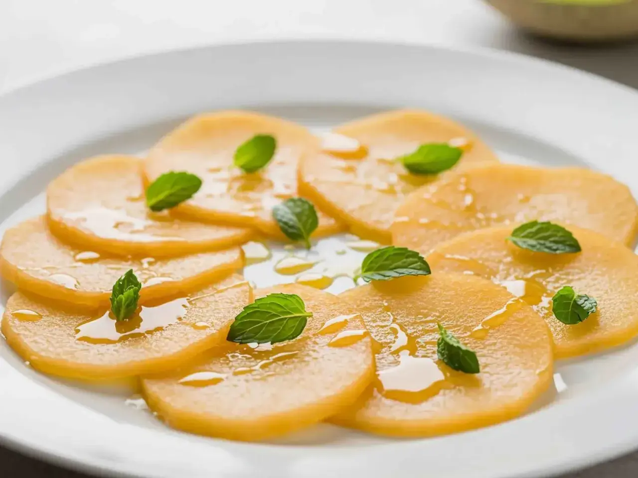 Carpaccio de melon frais, arrosé d'un filet de miel et garni de feuilles de menthe, sur une assiette blanche.