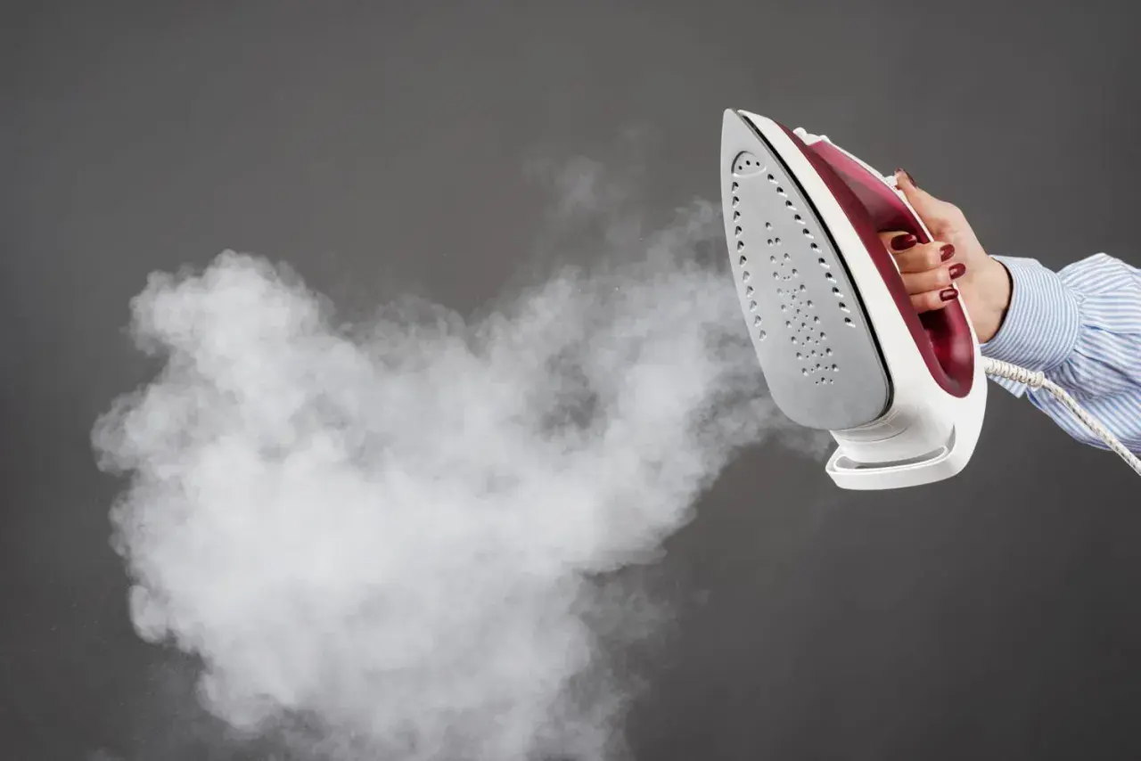 Une main tient un fer à repasser qui émet de la vapeur, suggérant l'action de détartrer le fer à repasser pour un entretien optimal.