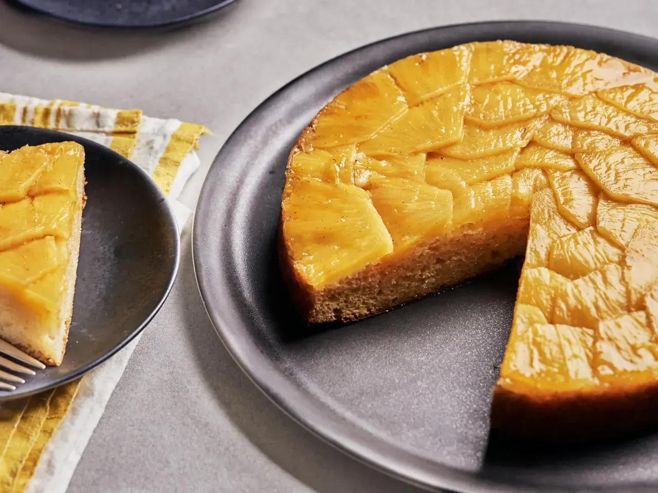 Un délicieux gâteau ananas renversé, avec une tranche servie sur une assiette noire. Les fruits caramélisés brillent sous la lumière.