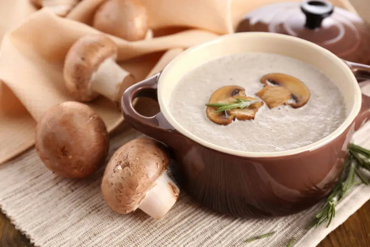 Un bol de soupe de champignons de Paris onctueuse, garnie de lamelles de champignons et de romarin. Des champignons frais entourent le plat.