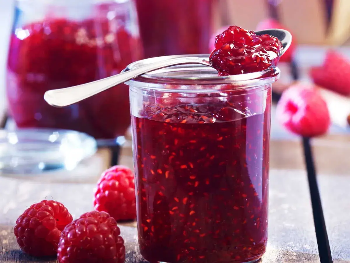 Un pot de confiture de framboise grand-mère, garni d'une cuillère pleine de cette délicieuse préparation. Des framboises fraîches entourent le pot.