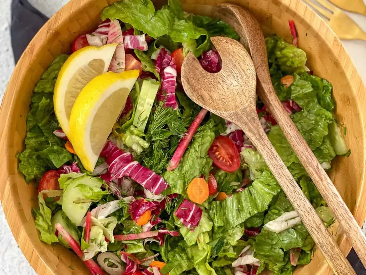 Salade verte fraîche et colorée, parfaite pour une recette légère. Radis, concombre, tomates cerises et citron dans un saladier en bois avec des couverts.
