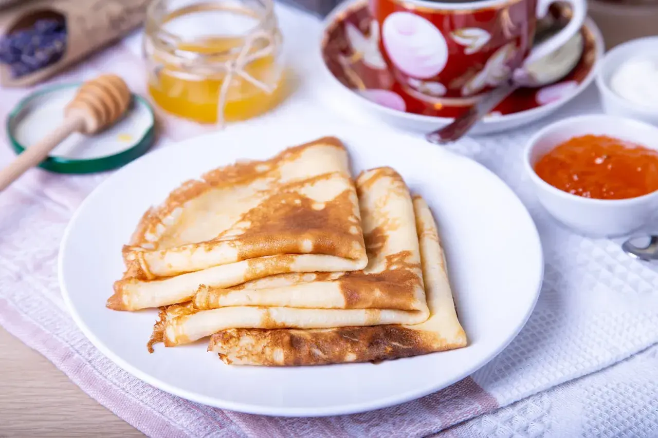 Pile de crêpes dorées, prêtes pour la préparation de la pâte à crêpes. Accompagnées de miel, confiture et d'une tasse de thé.