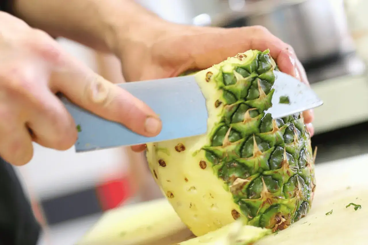 Mains expertes d&eacute;montrant comment couper un ananas, retirant la peau verte &eacute;pineuse avec un couteau bleu.