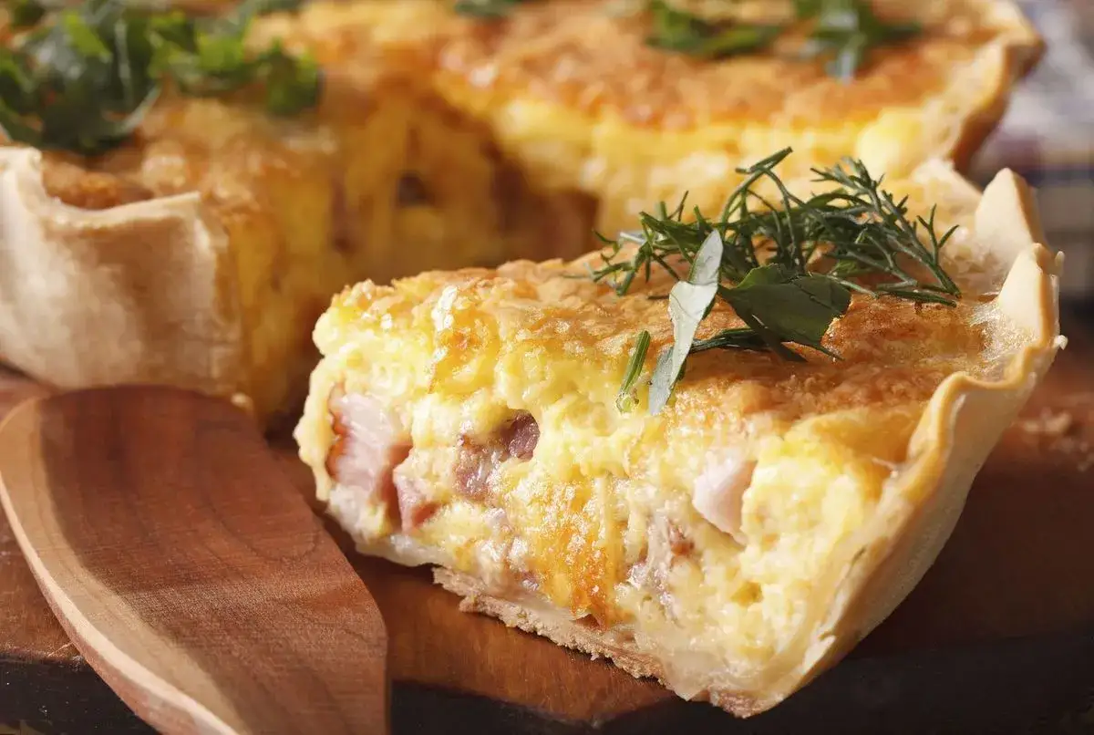 Une part de v&eacute;ritable quiche lorraine, garnie d'herbes fra&icirc;ches, sur une planche en bois avec une cuill&egrave;re.