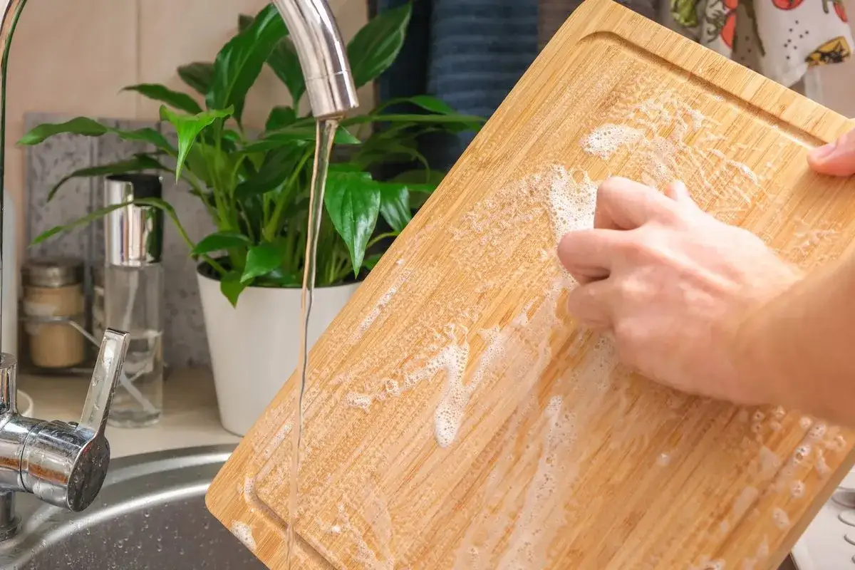 Nettoyer planche à découper en bois avec du bicarbonate de soude et de l'eau. Une main frotte la surface savonneuse sous le robinet.
