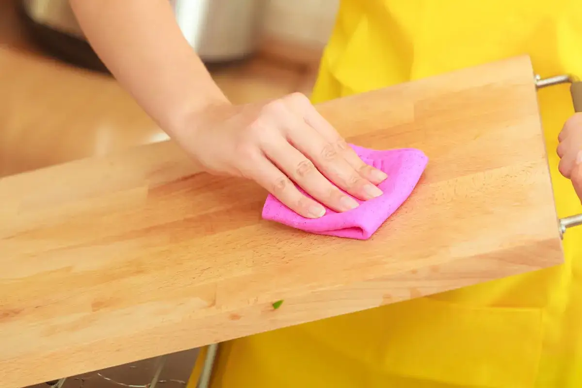 Main d'une personne nettoyant une planche &agrave; d&eacute;couper en bois avec un chiffon rose.