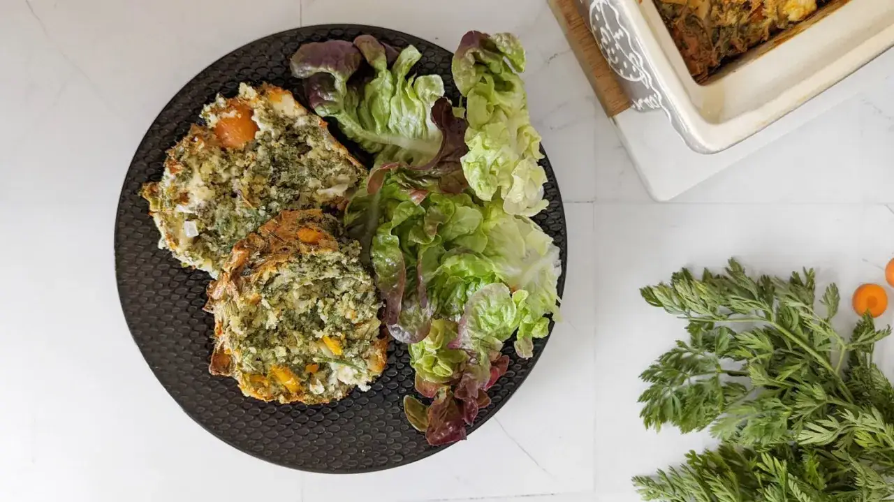 Deux parts de cake fanes de carottes, accompagn&eacute;es d'une salade verte fra&icirc;che. Des fanes de carottes brutes sont dispos&eacute;es &agrave; c&ocirc;t&eacute;.