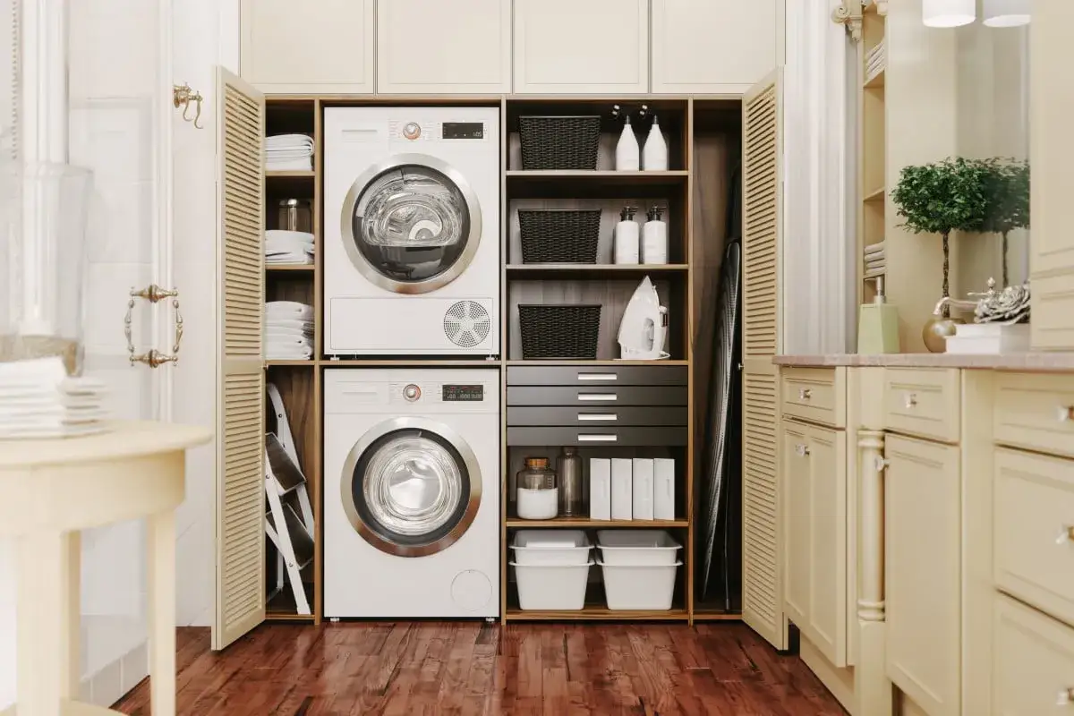 Un lave-linge et un s&egrave;che-linge superpos&eacute;s dans une buanderie bien rang&eacute;e, avec des paniers, des bouteilles et un fer &agrave; repasser sur des &eacute;tag&egrave;res.