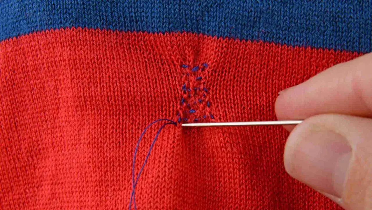 Main d'une personne utilisant une aiguille et du fil bleu pour comment recoudre un tissu d&eacute;chir&eacute; sur un v&ecirc;tement ray&eacute; rouge et bleu.