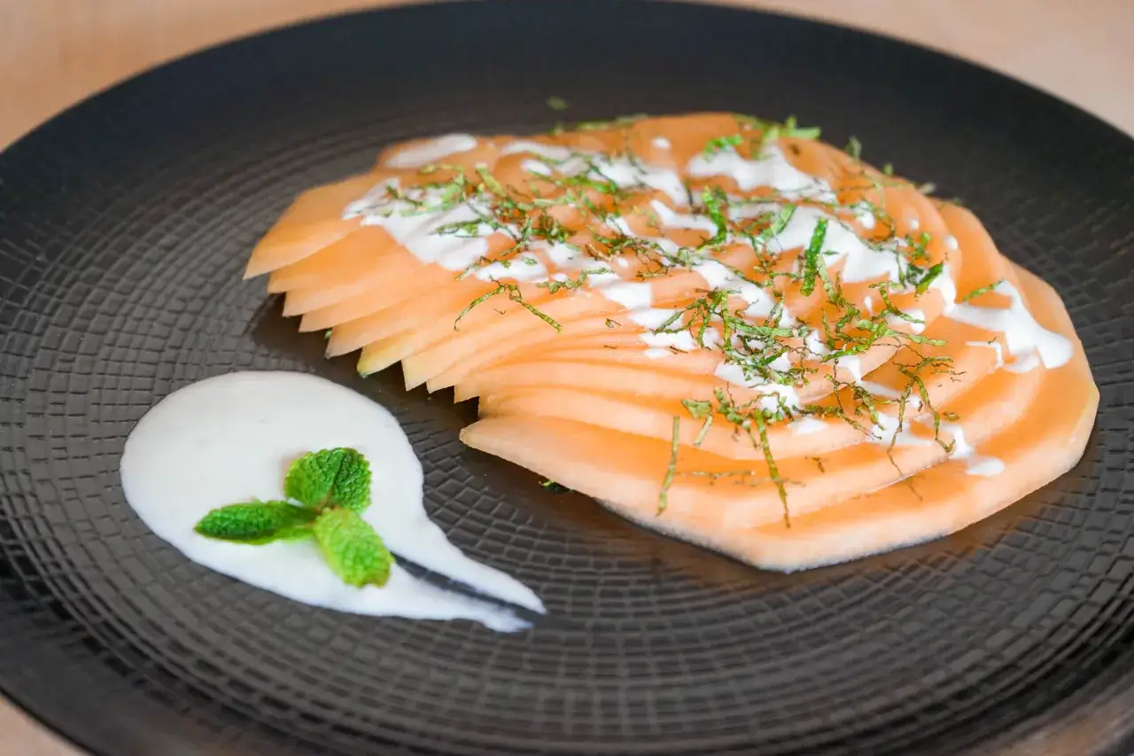 Délicieux carpaccio de melon, finement tranché, arrosé d'une sauce crémeuse et parsemé d'herbes fraîches. Servi avec une touche de crème et de menthe.