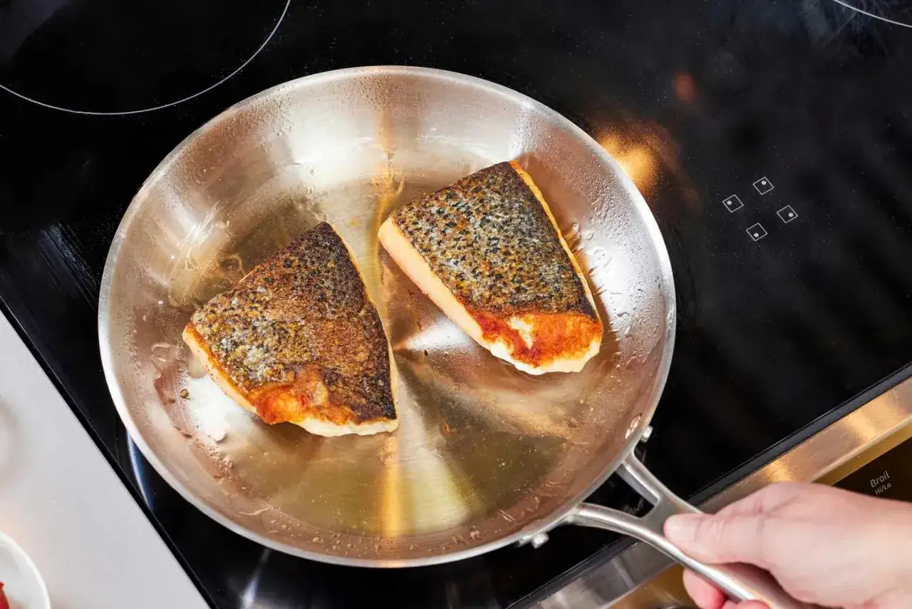 Deux pavés de saumon dorent dans une poêle en inox sur une plaque à induction. Attention, la poêle en inox peut devenir très chaude.